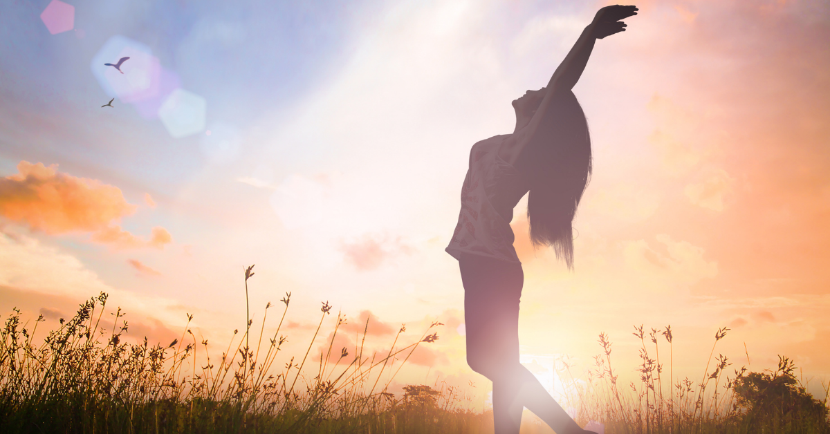 Woman acting freed of her burdens in a field with the sun setting in the background.