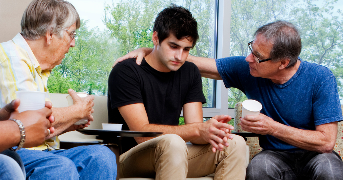 Young man sits in a chair while one older man has a hand around him and another older person speaks to him