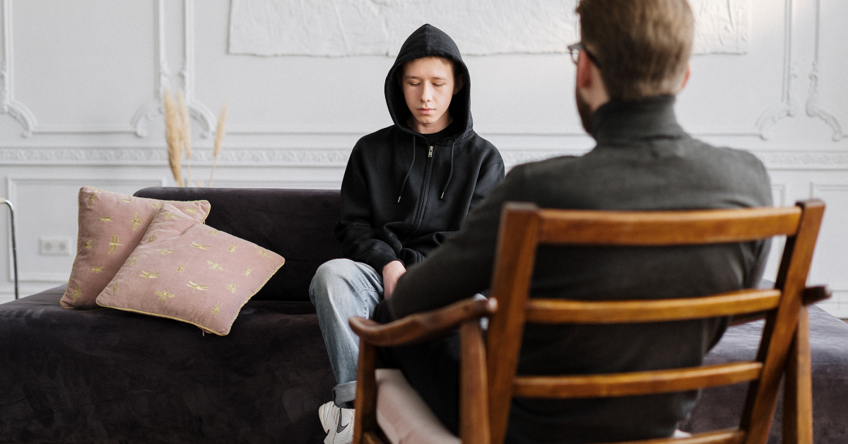 Young man in a hoodie sits on a couch while talking to a therapist whose face we cannot see