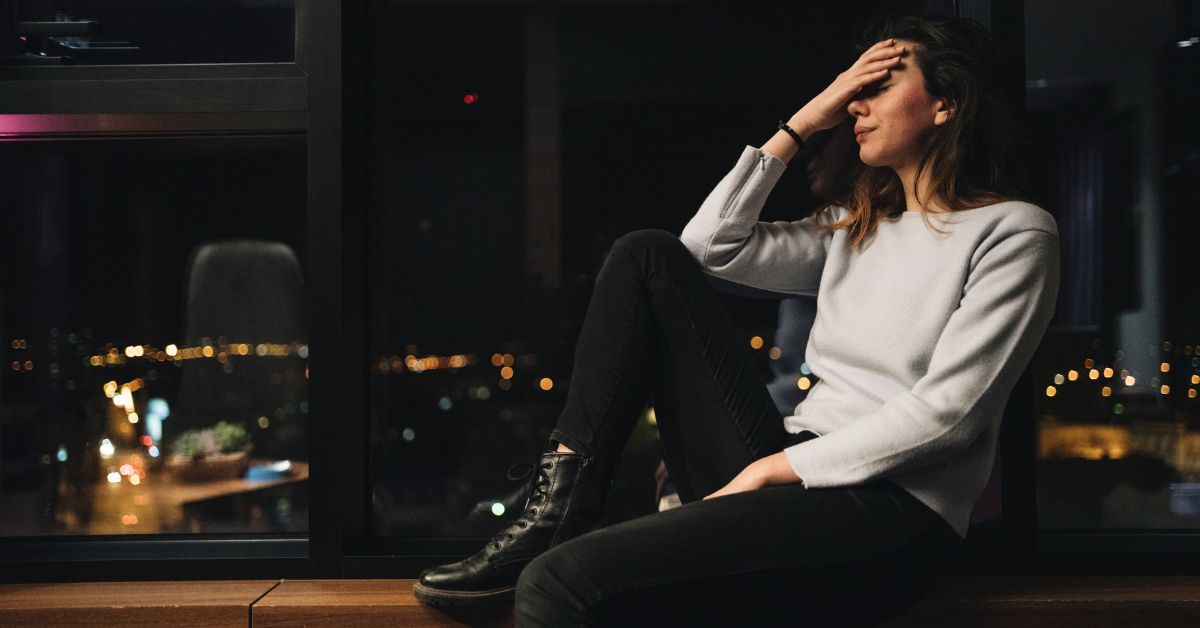 An adult woman sitting on a ledge , indoors, that is next to a window. She is clearly upset